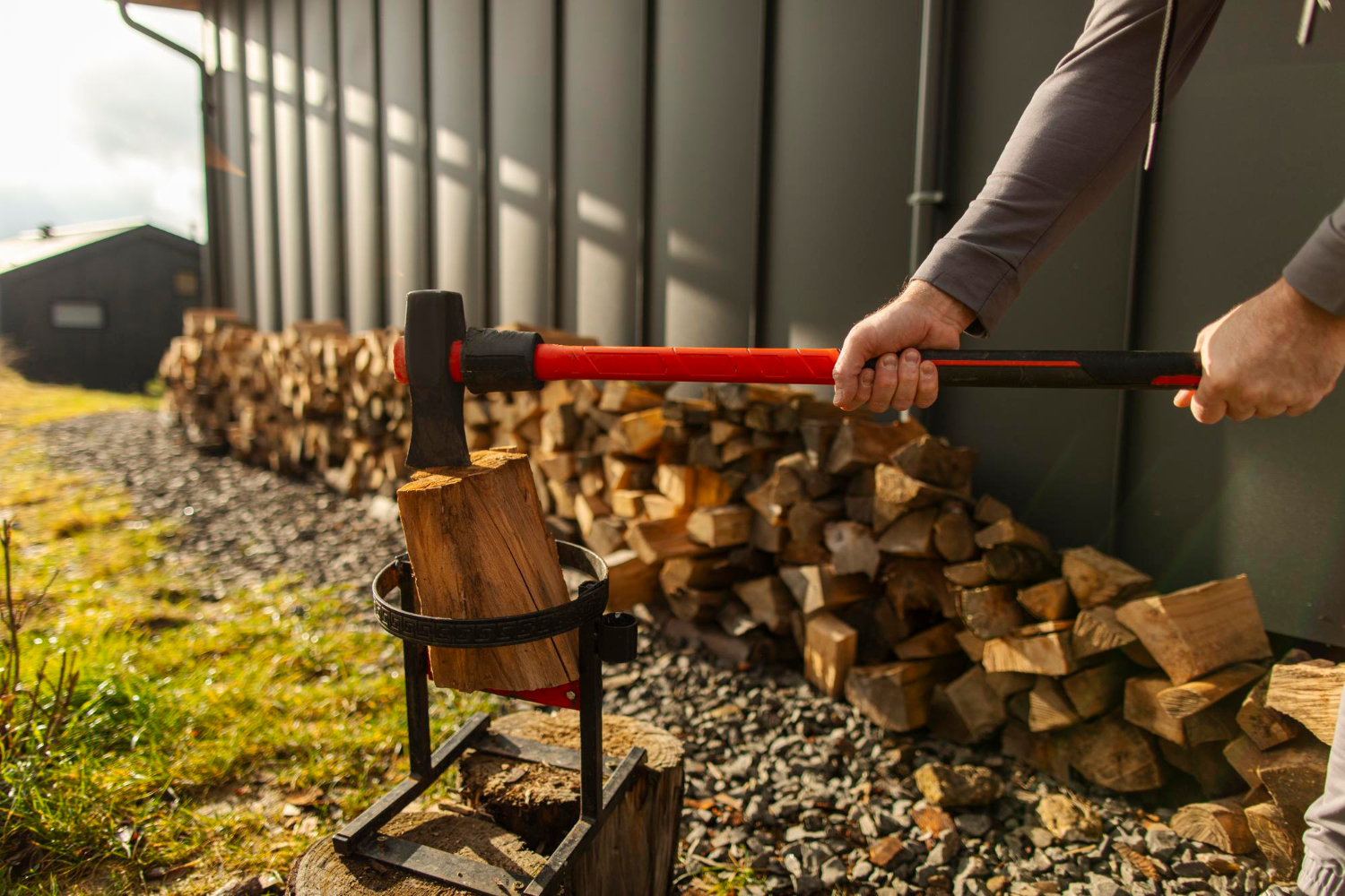 Homme utilisant une hache pour fendre un morceau de bois, debout près d'une pile de bois de chauffage bien rangée.